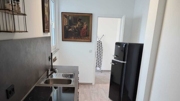 Une cuisine moderne dotée d’un évier en acier inoxydable, d’un four encastré et d’un réfrigérateur noir. Un tableau est accroché au mur et une serviette à motifs est suspendue à une porte.