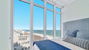 Ein Schlafzimmer mit großem Fenster mit Blick auf Strand und Meer, einem Bett mit blauer Bettdecke und einem grauen Kopfteil.