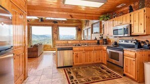 A wooden cabin kitchen with a stone-tiled floor, wooden cabinets, a stove, and a view of the mountains through large windows.