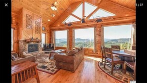 A wooden cabin with a stone fireplace, a sofa, a dining table, and large windows offering a mountain view.