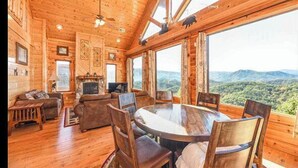 A wooden cabin interior with a fireplace, a round dining table, and a view of the mountains.