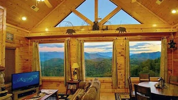 A wooden cabin interior with a large window offering a mountain view, a television, and a dining area.