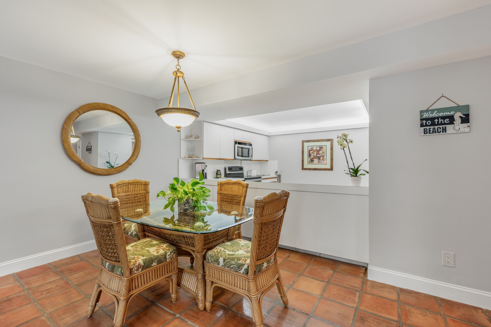 Un coin repas avec une table à plateau en verre, des chaises en osier, un luminaire suspendu et une cuisine en arrière-plan.
