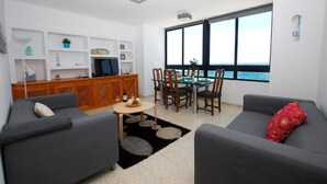 Una sala moderna con dos sofás grises, una mesa de comedor con sillas, un televisor en un mueble de madera y un ventanal con vistas al mar.