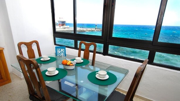 Comedor con mesa de vidrio, cuatro sillas y vista al océano a través de amplias ventanas.