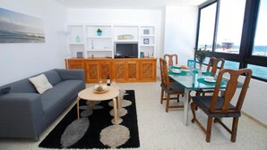 Una sala de estar moderna con sofá, mesa de centro y comedor con sillas. Hay un televisor sobre un mueble de madera, y un ventanal con vistas al mar.