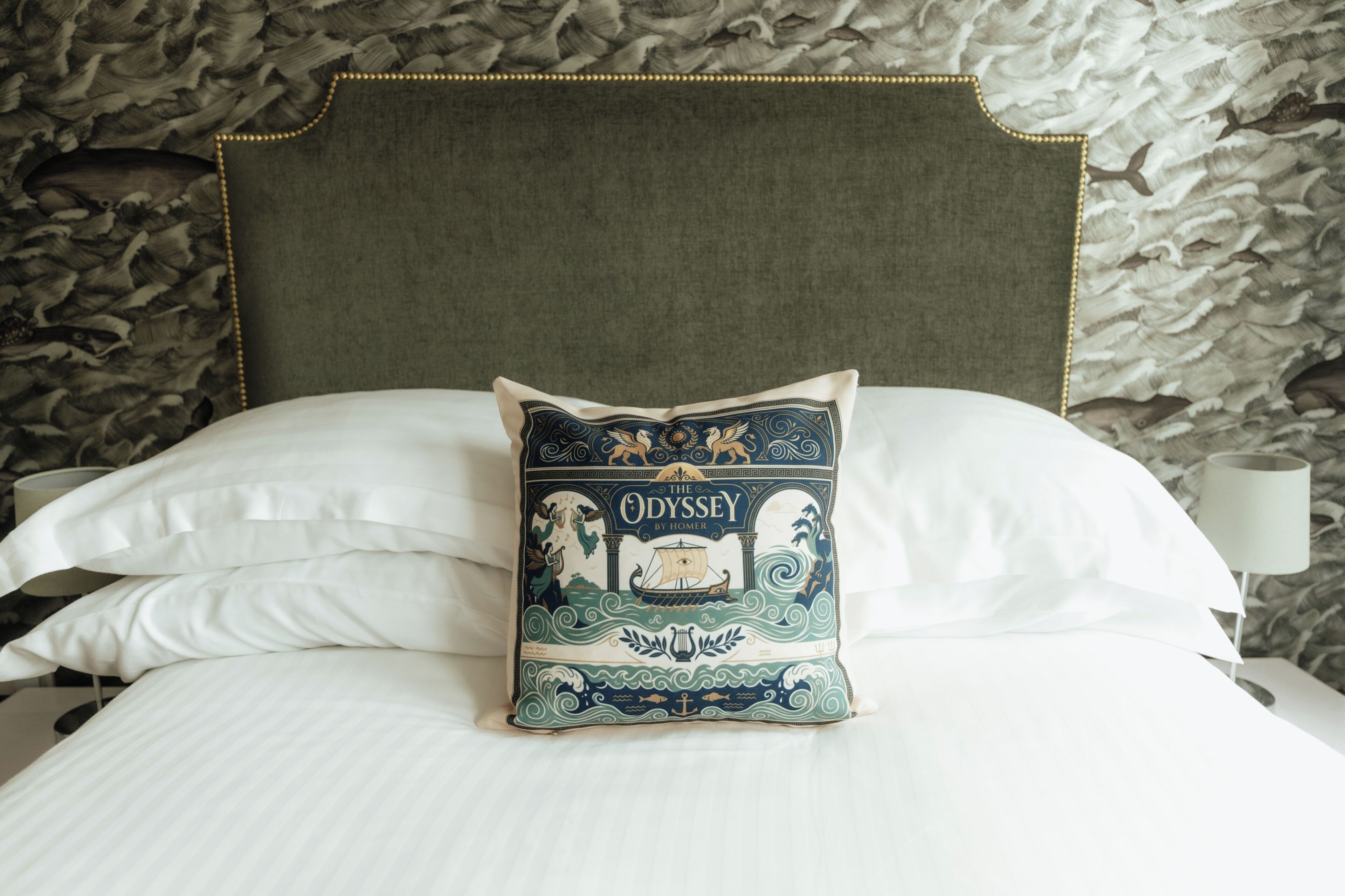 A bed with a decorative pillow featuring a nautical theme, a plain headboard, and a patterned wall behind.