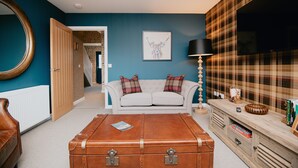 A living room with a large blue wall, a white sofa with plaid cushions, a wooden coffee table, a brown trunk, a plaid-patterned TV stand, and a mounted TV. - The Seafield Arms Hotel Cullen – Self Catering (Buckie)
