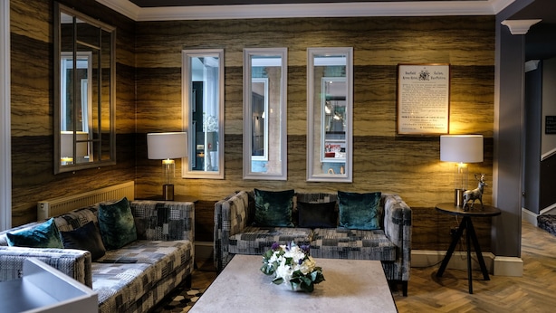 A cozy living room with a patterned sofa, a coffee table with a floral centerpiece, and a wall-mounted mirror. - The Seafield Arms Hotel Cullen – Self Catering (Buckie)