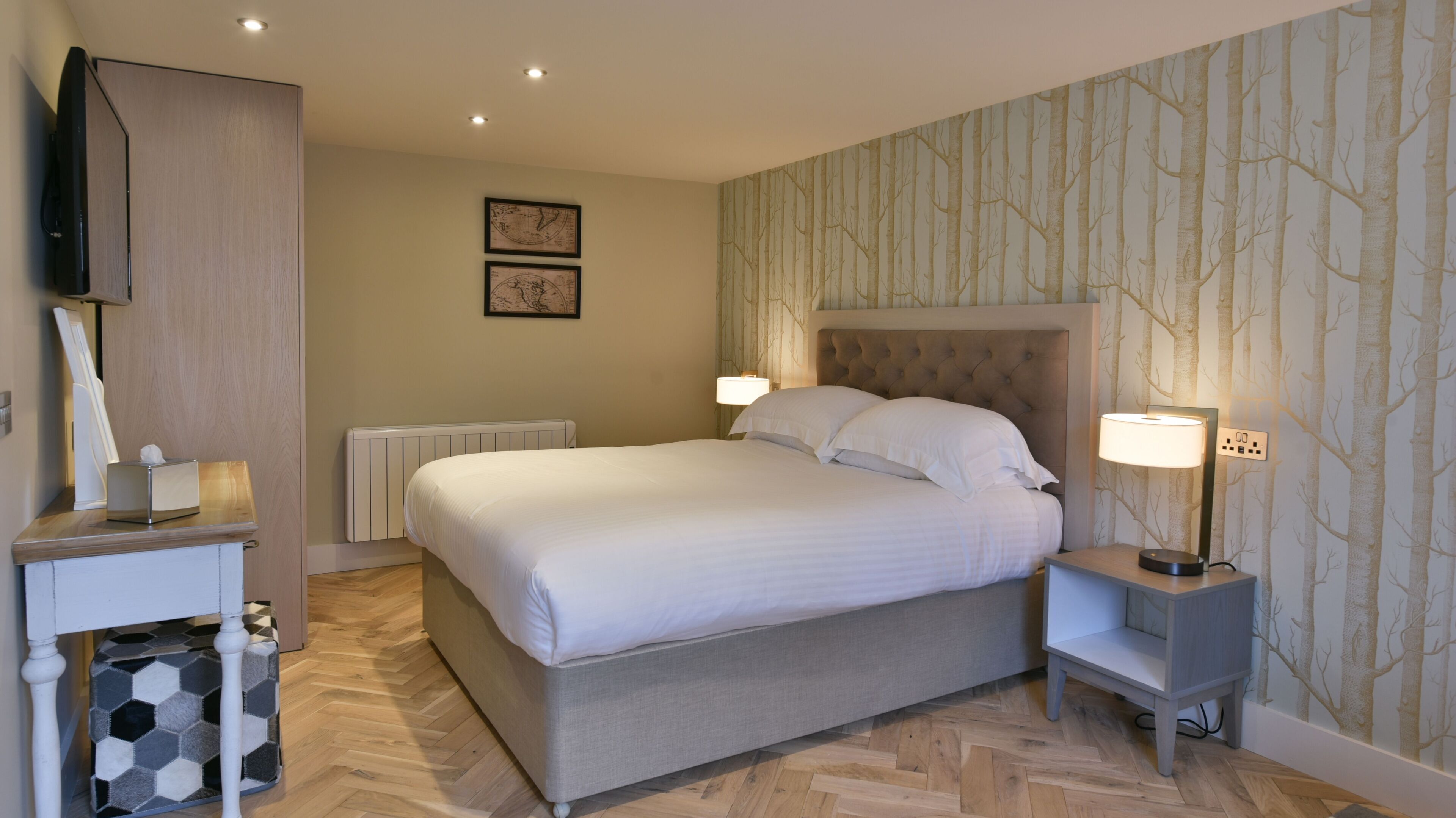 A neatly arranged hotel room with a large bed, two bedside lamps, a flat-screen TV, a small table with a tissue box, and a patterned wallpaper.