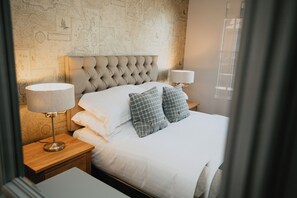 Una cama bien tendida con sábanas blancas y dos almohadones cuadriculados, flanqueada por dos lámparas de noche sobre mesitas de madera.