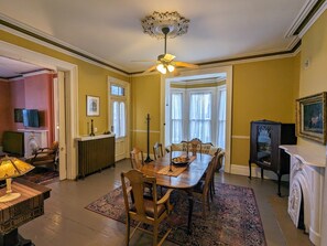 Une salle à manger avec une table en bois, des chaises, un lustre et une grande fenêtre en saillie agrémentée de rideaux.
