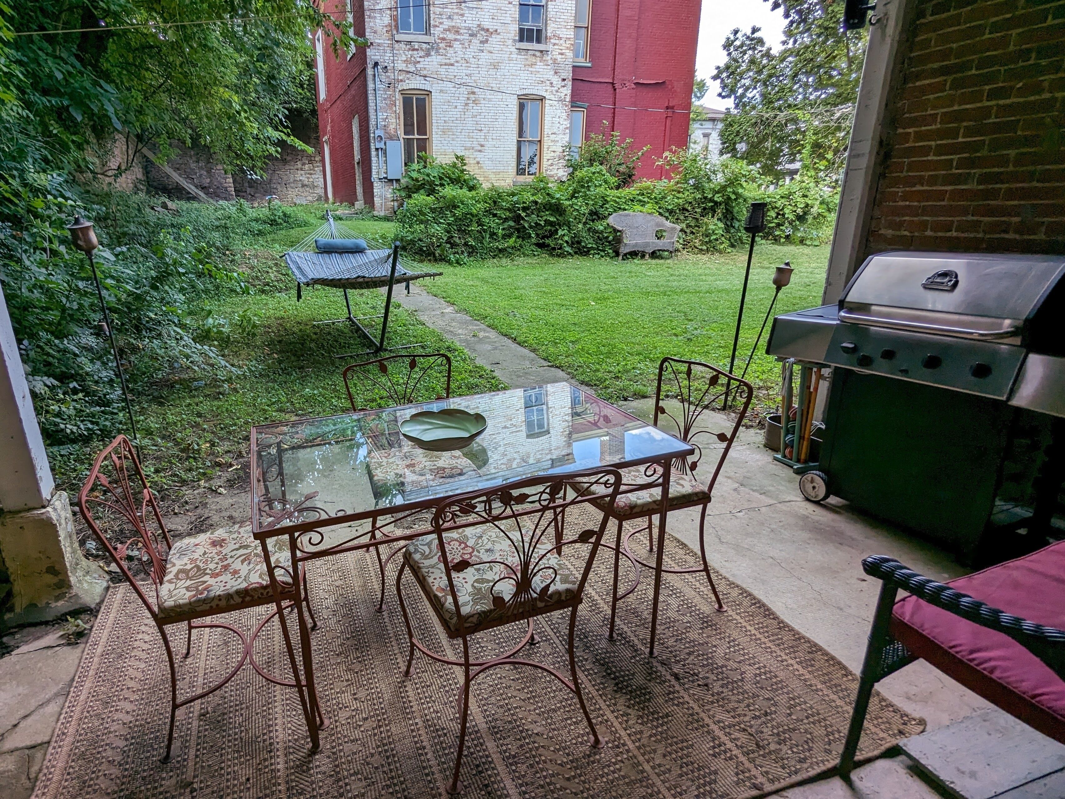 Une terrasse extérieure avec une table en verre et des chaises en métal, un barbecue et un petit jardin avec une pelouse et un sentier.