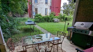 Eine Terrassenausstattung im Freien mit Glastisch, Metallstühlen, einem Grill und einem kleinen Gartenbereich mit Rasen und einem Weg.