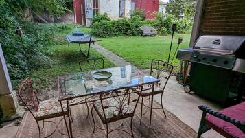 An outdoor patio with a glass table and metal chairs, a grill, and a small garden area with a lawn and a pathway.