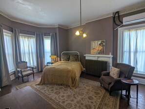 A bedroom with a bed, a chair, a table, a radiator, and a painting on the wall.