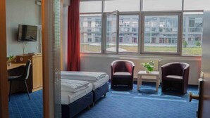 A hotel room with a large bed, two armchairs, a small table with a plant, a desk with a TV, and a window with a view of buildings.