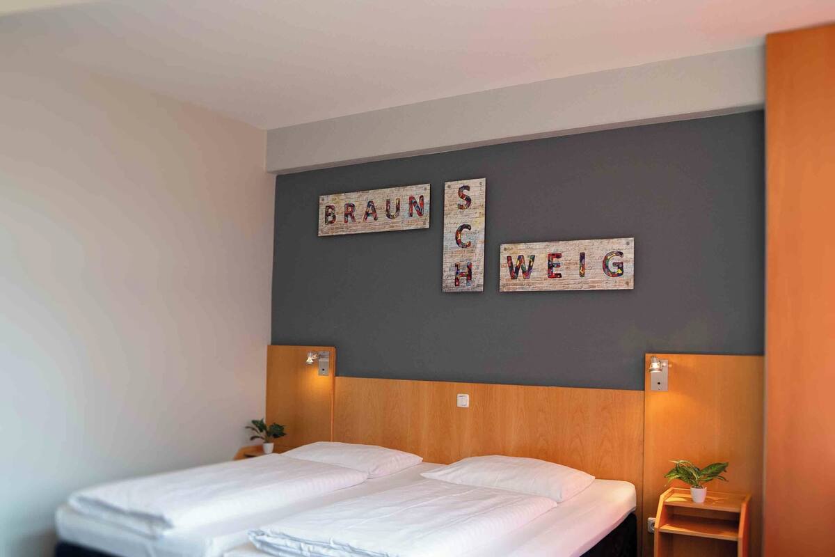 A hotel room with two beds, a wooden headboard, a small bedside table with a plant, and a wall with the words "BRAUN SCH WEIG" displayed.