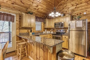 A wooden cabin kitchen with granite countertops, wooden cabinets, a stainless steel refrigerator, and a hanging light fixture. - 2 King Bedroom Cabin with Private Hot Tub, Pool Table & Arcade (SEVIERVILLE)