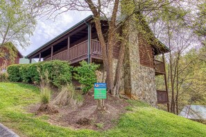 A cabin with a wooden deck and stone walls, surrounded by trees and greenery. - 2 King Bedroom Cabin with Private Hot Tub, Pool Table & Arcade (SEVIERVILLE)