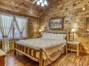 Une chambre à coucher dans une cabane en bois, avec un lit, deux tables de chevet, une petite table, une lampe, un tableau et une fenêtre avec des rideaux.