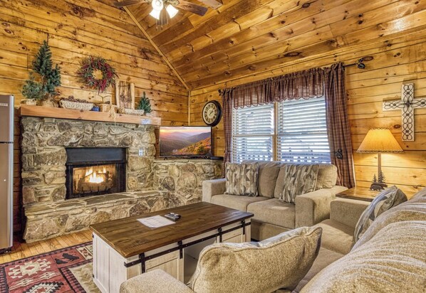 A cozy cabin interior with a stone fireplace, wooden walls, a sofa, and a television. - 2 King Bedroom Cabin with Private Hot Tub, Pool Table & Arcade (SEVIERVILLE)