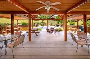 Une terrasse extérieure avec un plancher en bois, des tables et des chaises en métal, un ventilateur de plafond et une vue sur une piscine et un espace verdoyant.