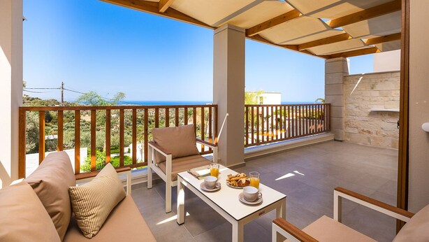 Ein Balkon mit Holzgeländer, einer Couch, einem Tisch mit Frühstücksgebäck und Blick auf das Meer.