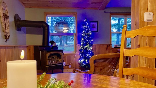 A cozy room with a wooden table, a lit candle, a Christmas tree, and a wood-burning stove. - The Quiet Spot Cabin (Naples)