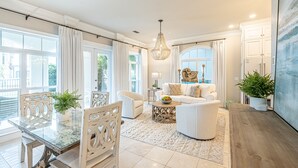 Un amplio salón con una mesa de comedor de cristal, sofás blancos y una araña.