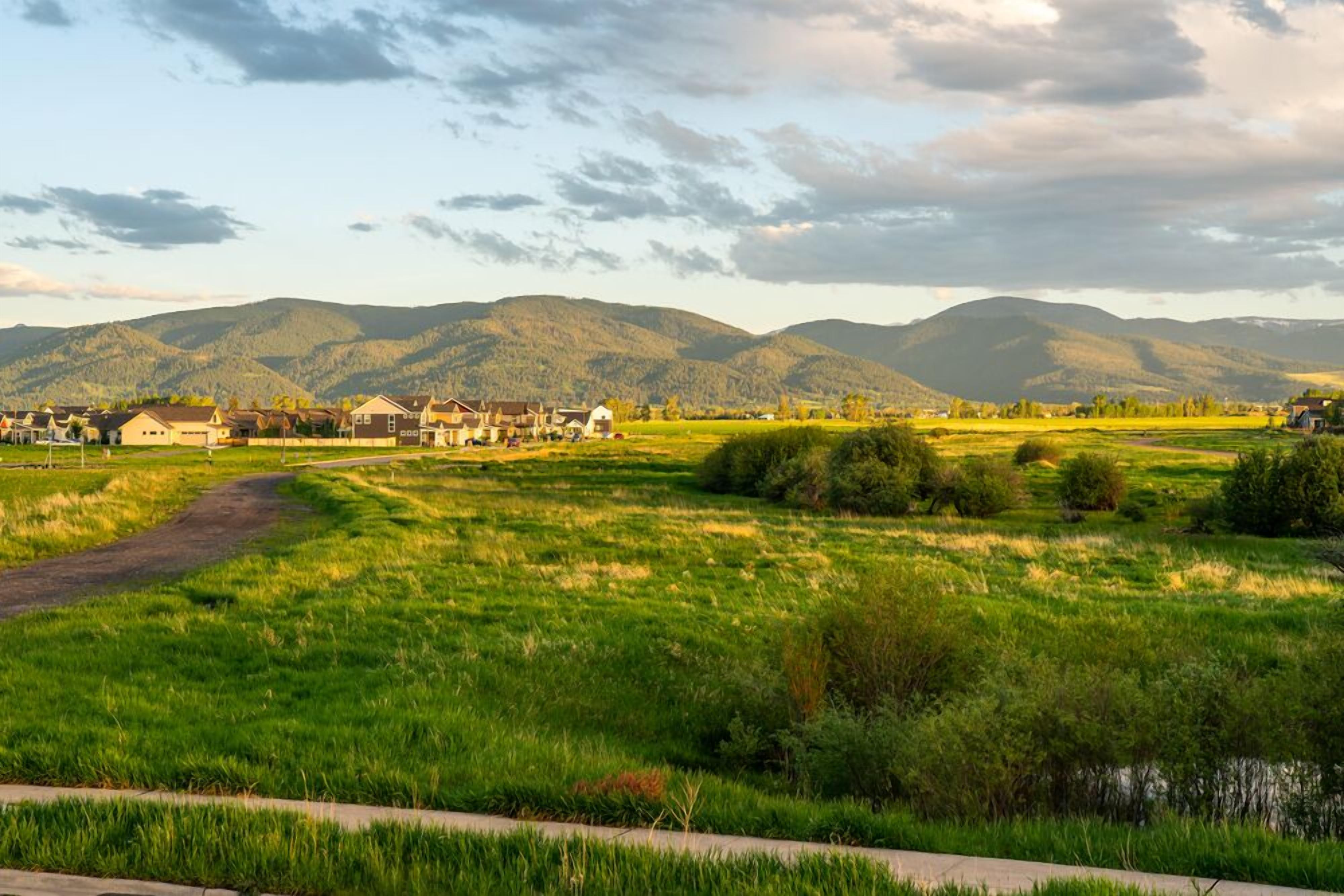 Un paysage rural avec un chemin de terre menant à une rangée de maisons, entouré de champs verdoyants et de montagnes en arrière-plan.