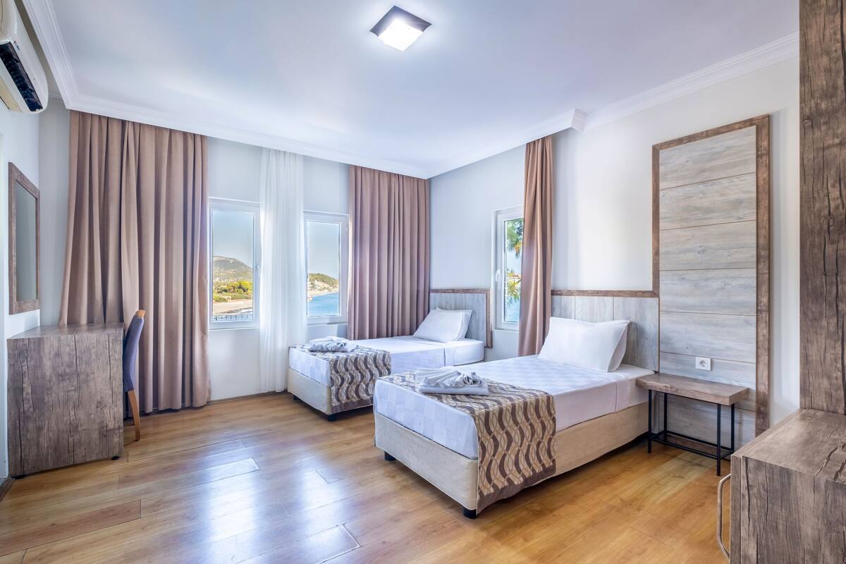A hotel room with two beds, a wooden floor, a large window with curtains, and a view of the outdoors.