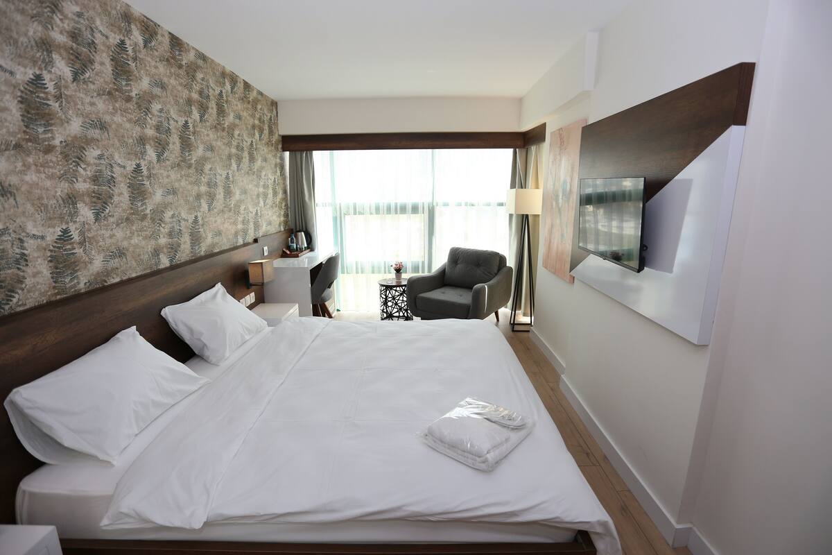 A hotel room with a large bed, a desk, a chair, a TV mounted on the wall, and a window with curtains.