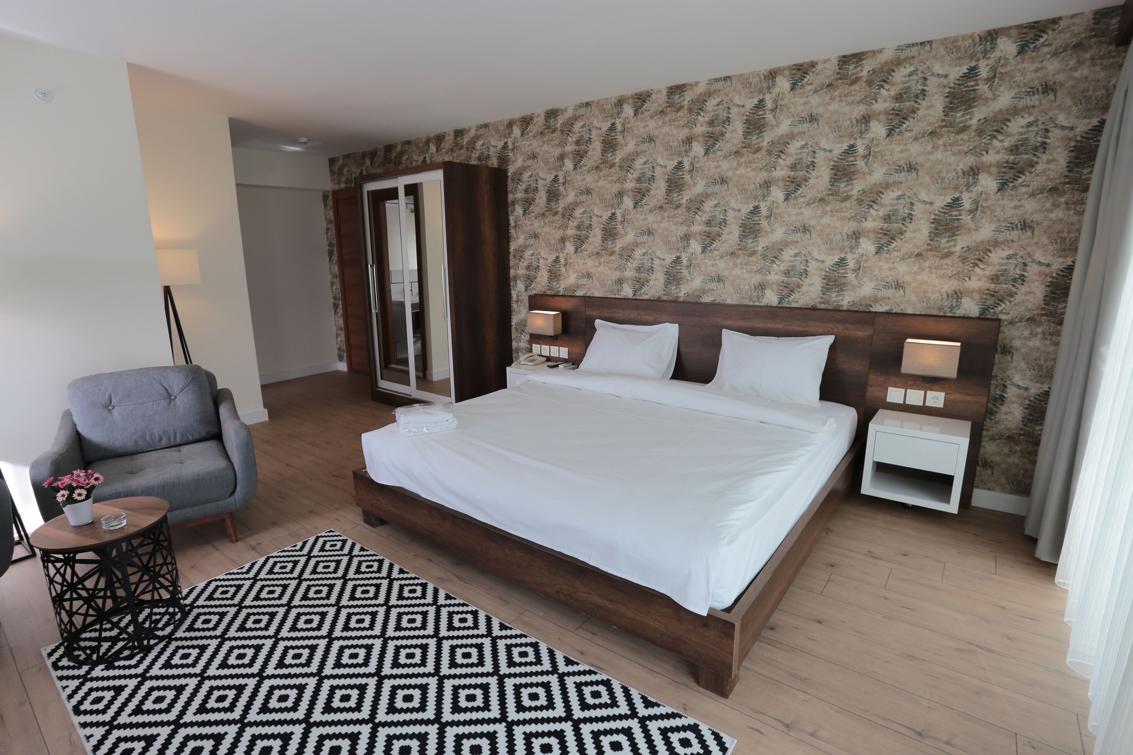 Una habitación de hotel con una cama grande, una silla, una mesita con un florero y una alfombra estampada.