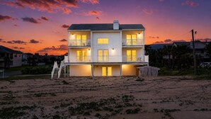 A two-story beachfront house with balconies, a wooden deck, and a sandy beach in the foreground.