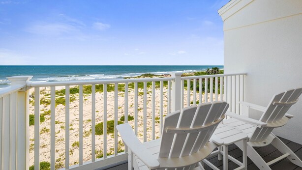 A balcony with white railings overlooking a beach with sand and sparse vegetation.