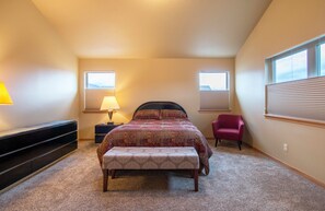 Un dormitorio con cama, banca, mesita de noche, lámpara, silla y ventanas.