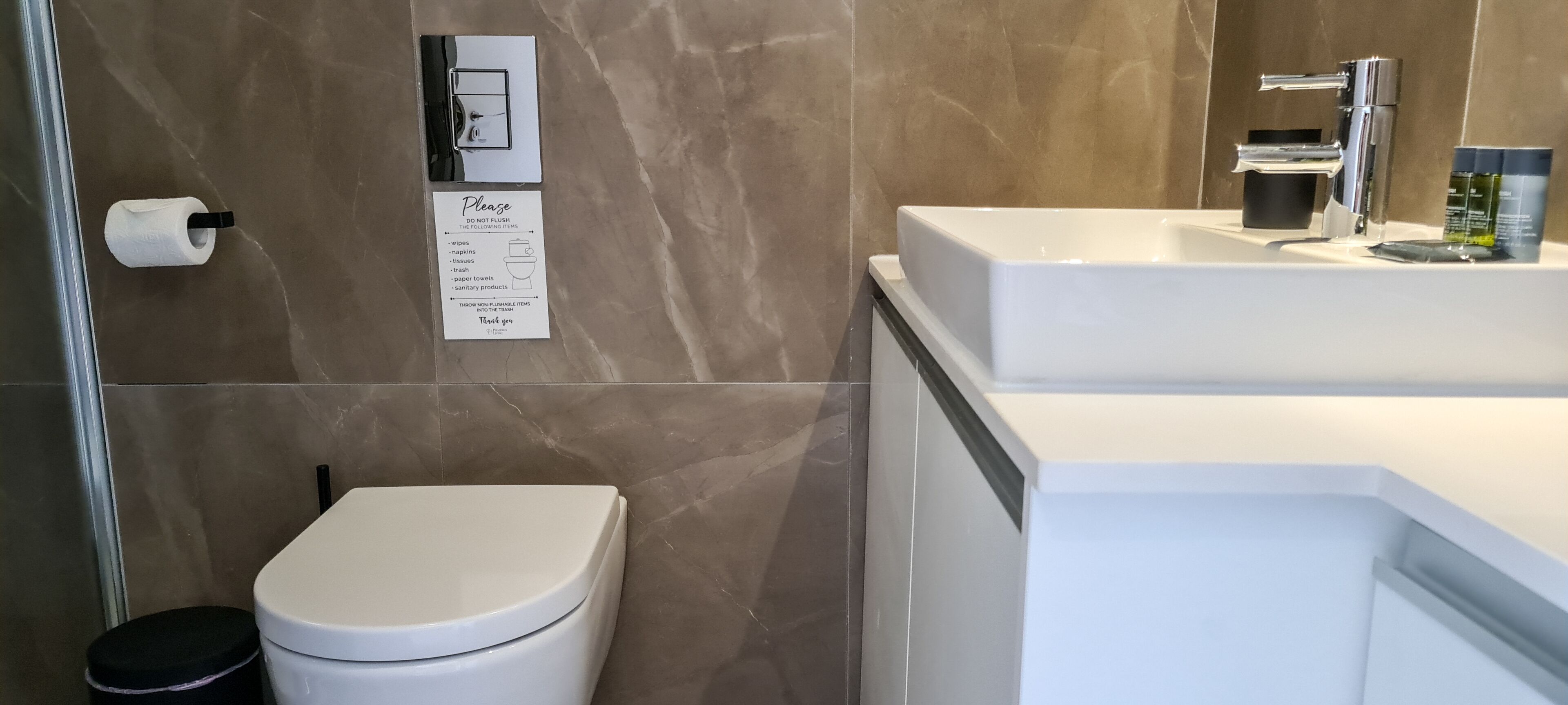 A modern bathroom with a toilet, sink, and a hand dryer. A sign instructs not to flush certain items.