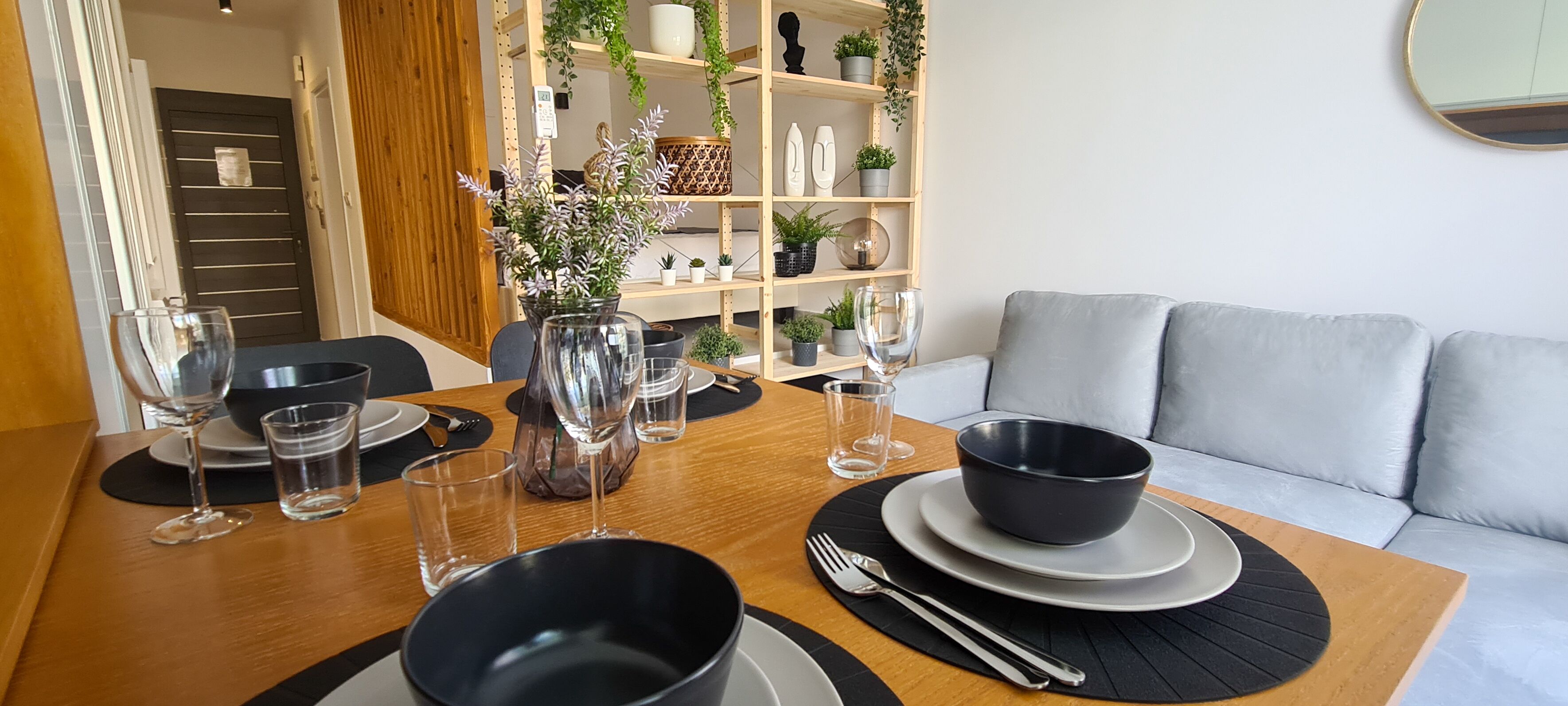 Een eetgedeelte met een houten tafel voor twee personen, voorzien van zwart-wit borden, kommen en glazen. Op de tafel staat een vaas met bloemen, en op de achtergrond een grijze bank.