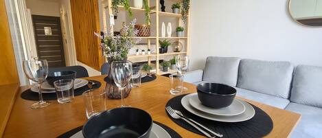 A dining area with a wooden table set for two, featuring black and white plates, bowls, and clear glasses. A vase with flowers is on the table, and a grey sofa is in the background.