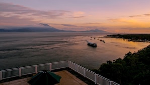 Vista costeira com um barco na água, uma montanha ao longe e um corrimão branco em primeiro plano.