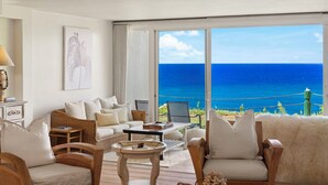 A spacious living room with a large window overlooking the ocean, furnished with wicker sofas, a coffee table, and a lamp. - Mahina Kai, Princeville Oceanfront Luxury, Steps to Hideaways (Princeville)