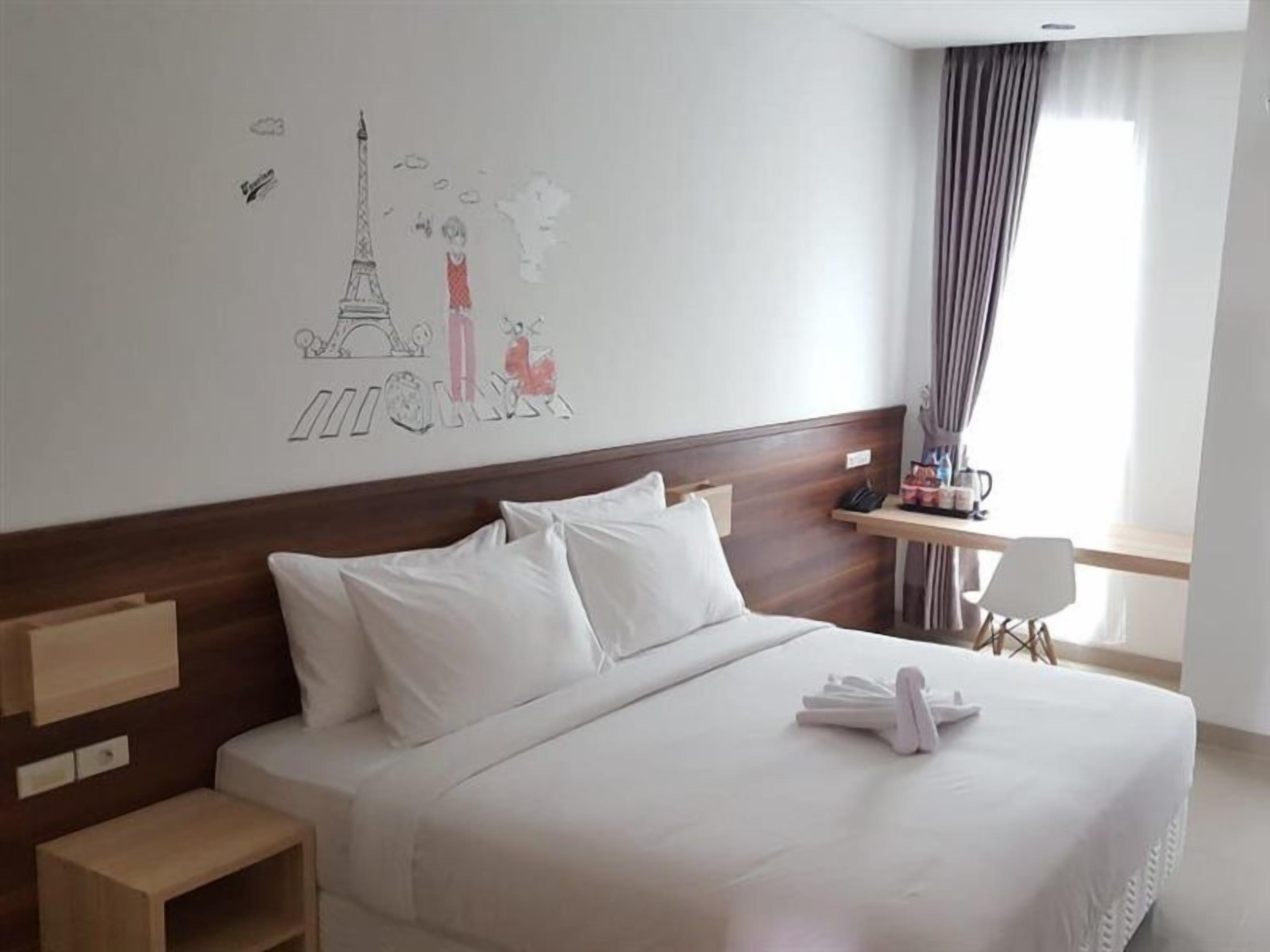 A hotel room with a bed, a desk, a chair, and a wall with a drawing.