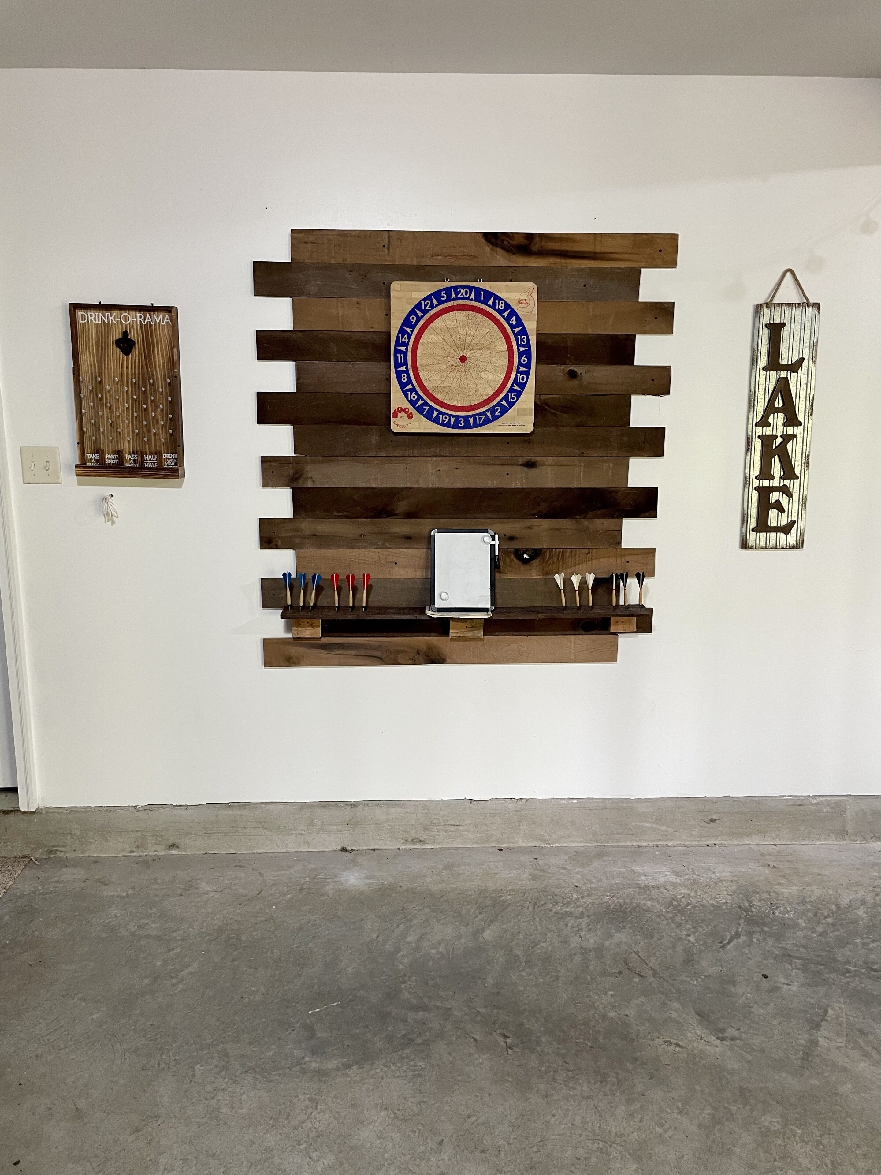 Un mur avec un jeu de fléchettes, une plaque en bois sur laquelle est écrit le mot « LAKE », et une étagère en bois contenant divers objets.