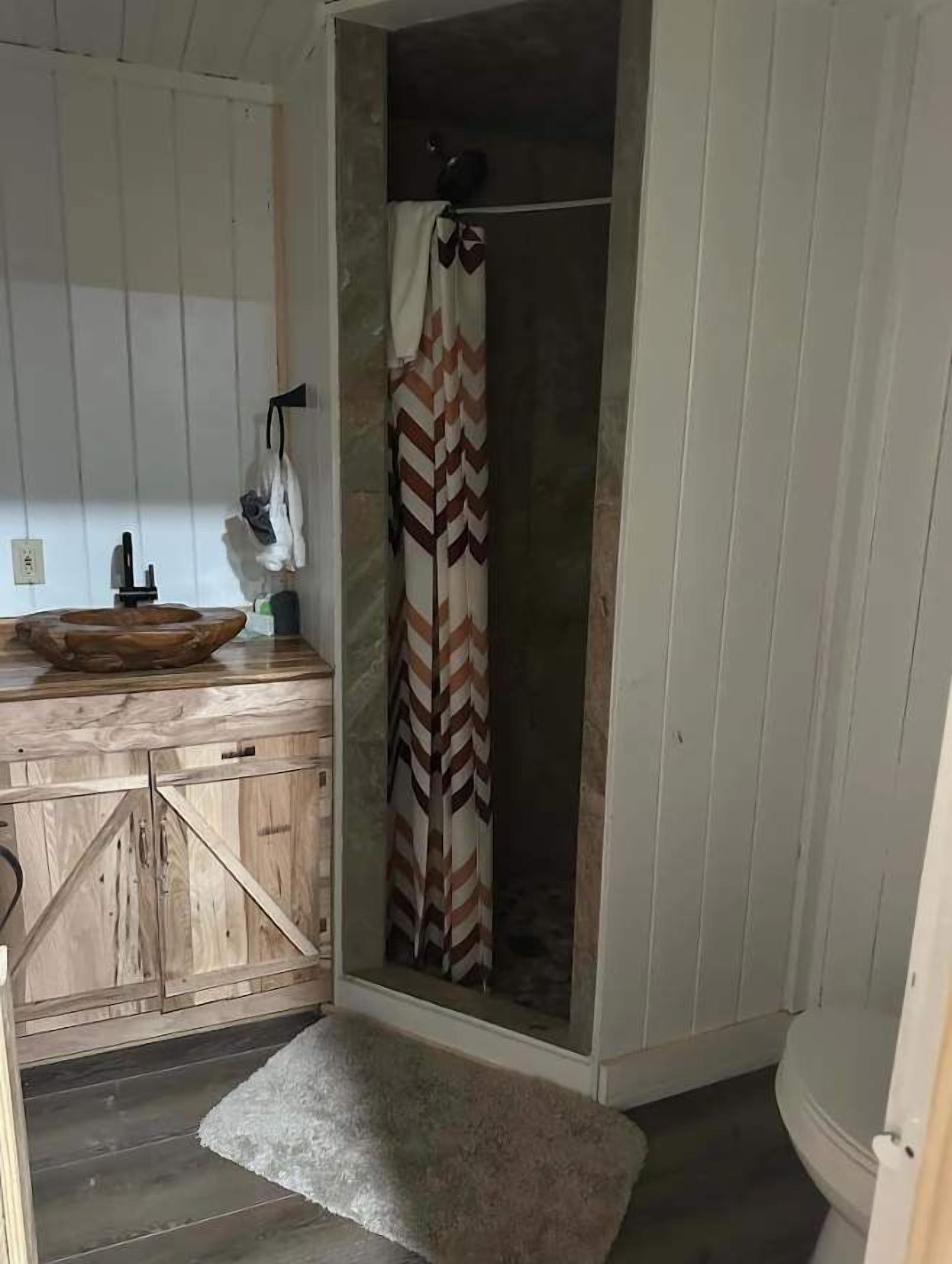 A bathroom with a wooden vanity, a shower with a patterned curtain, and a small rug on the floor.