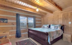 A wooden cabin bedroom with a bed, a nightstand, a chair, and a window with blinds.