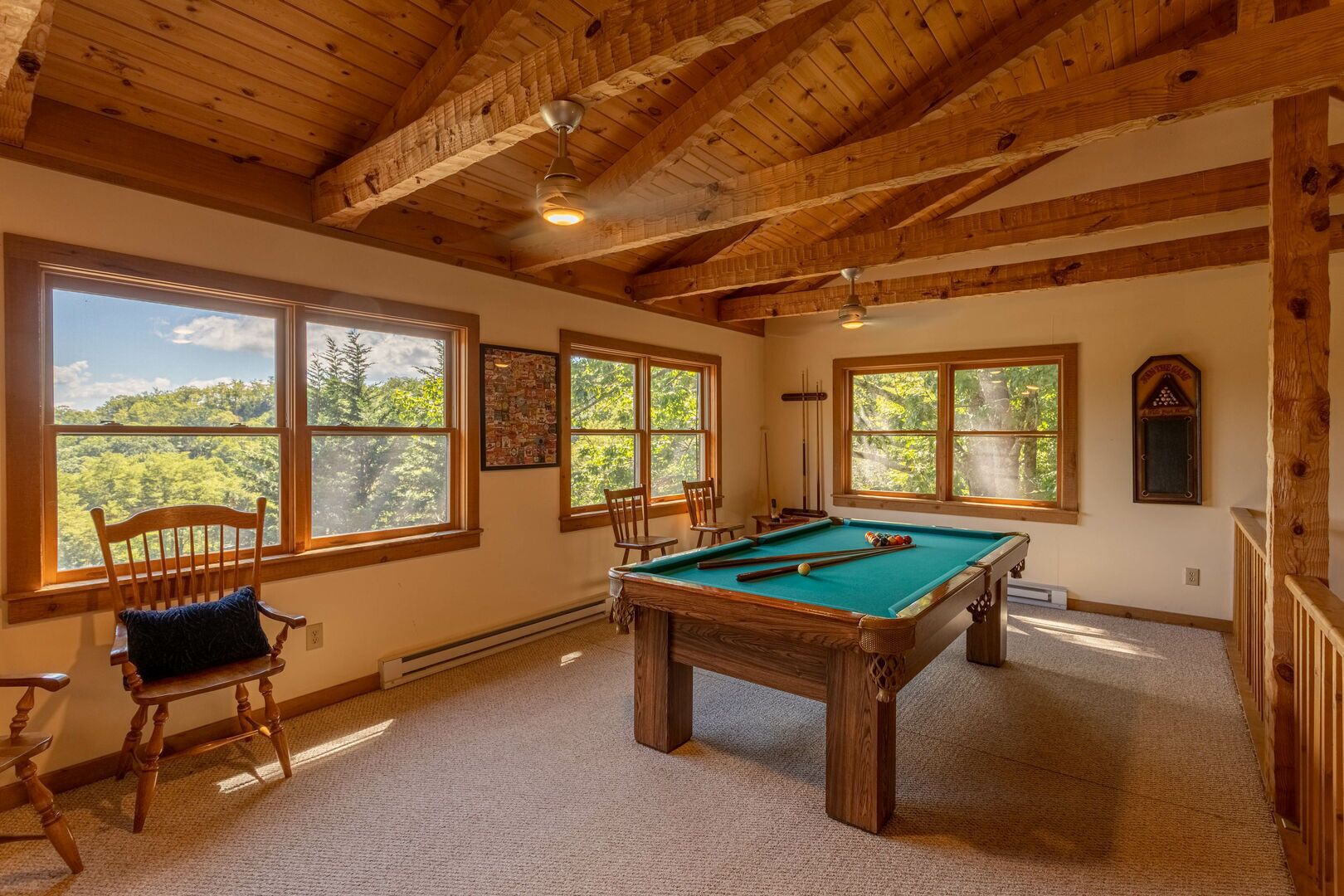 A pool table in a room with wooden beams and large windows overlooking a forest.