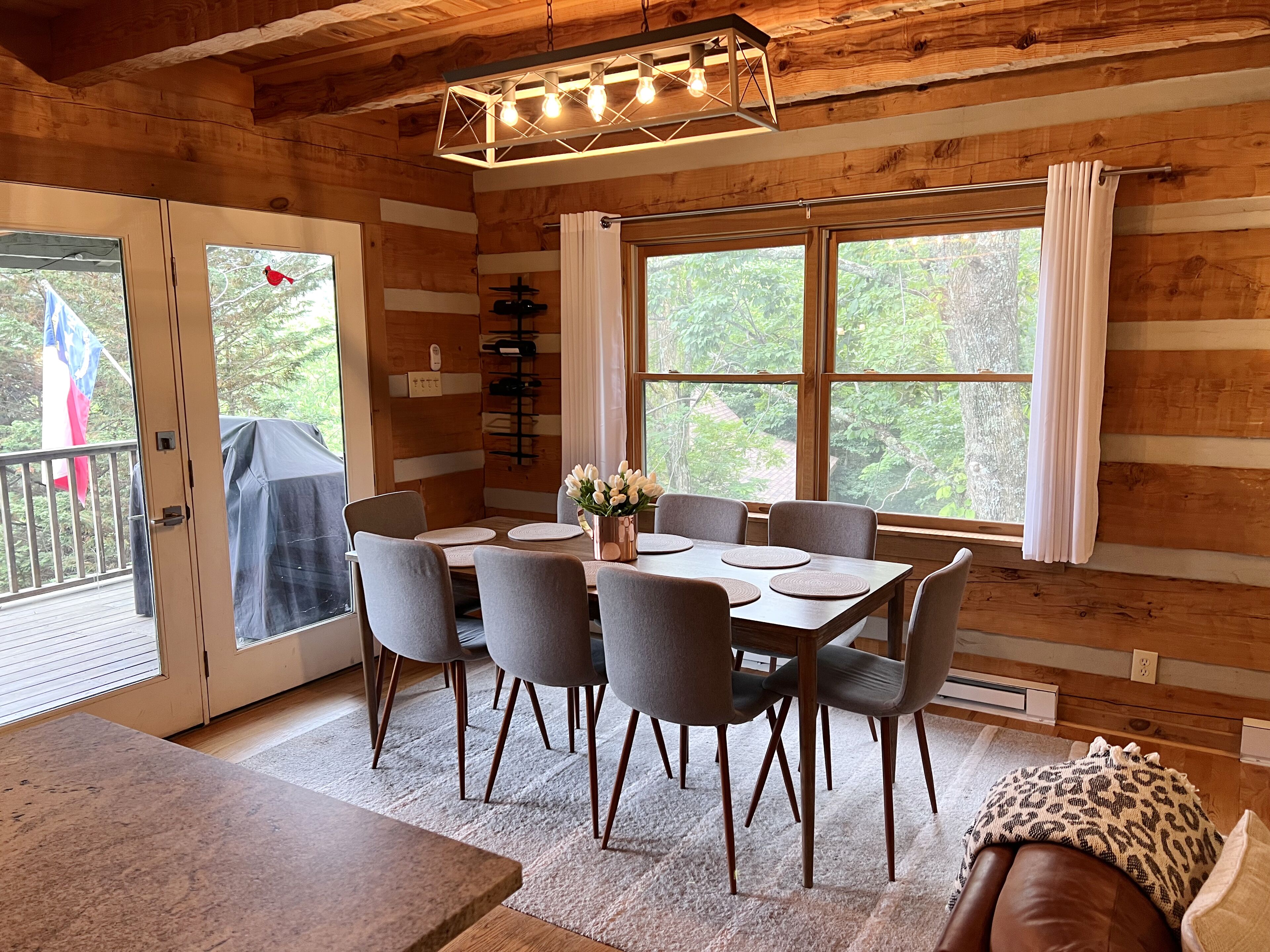 A wooden cabin interior with a dining area, large windows, and a ceiling light fixture.