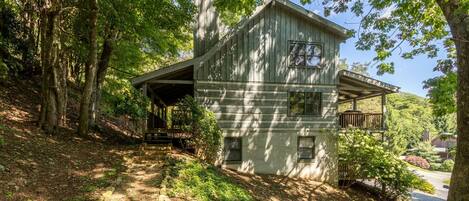 A two-story wooden cabin with a porch, surrounded by trees and a gravel path.