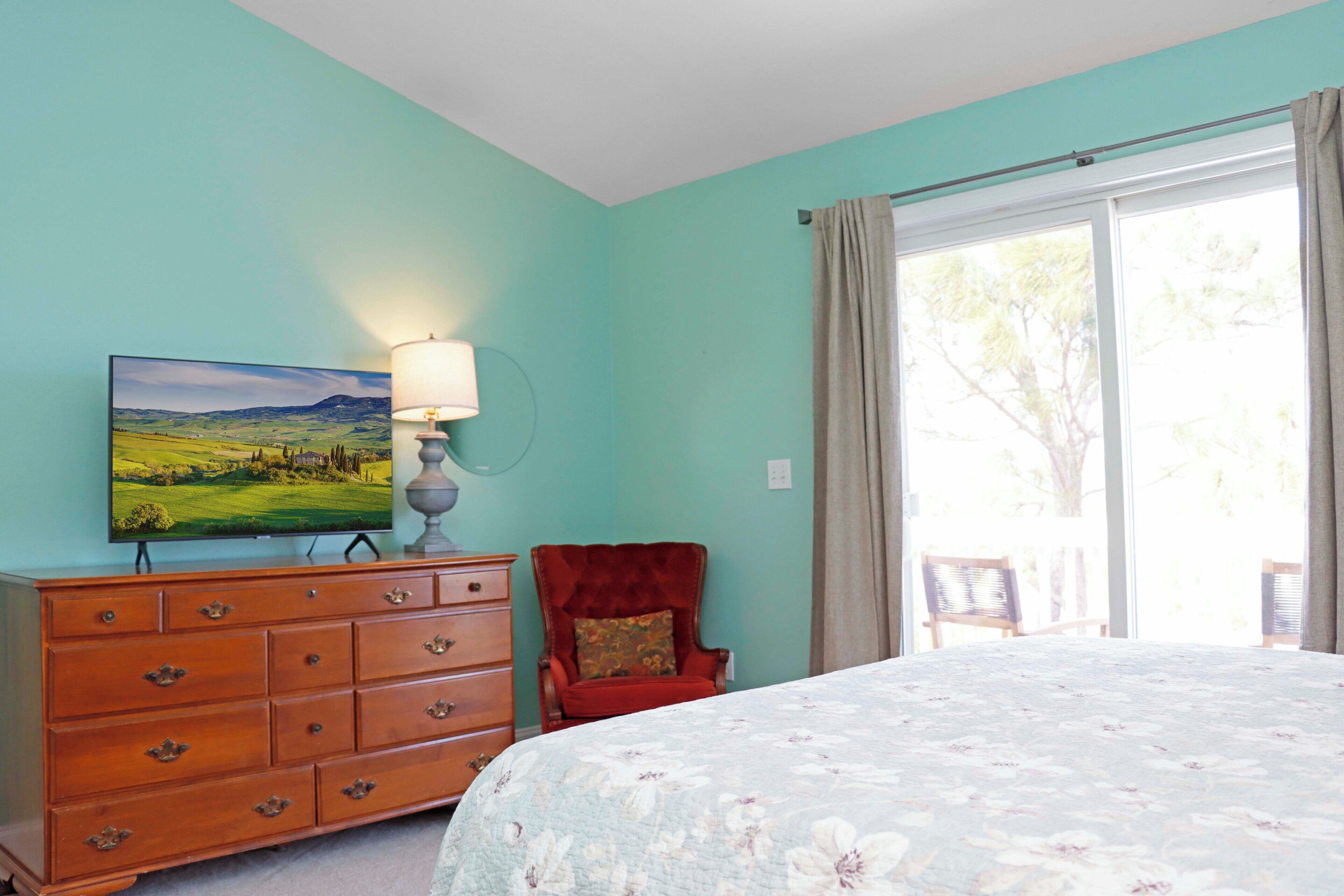 A bedroom with a bed, a dresser, a chair, a lamp, a television, and a window with curtains.